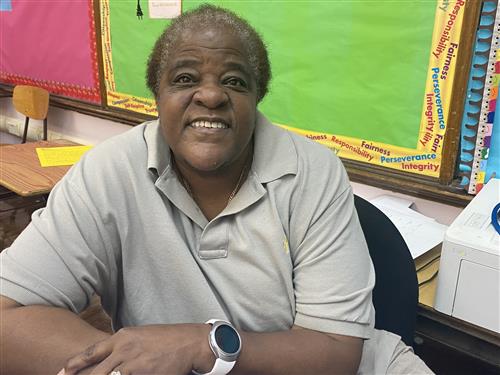 Teacher wearing tan shirt, smiling with watch on their wrist
